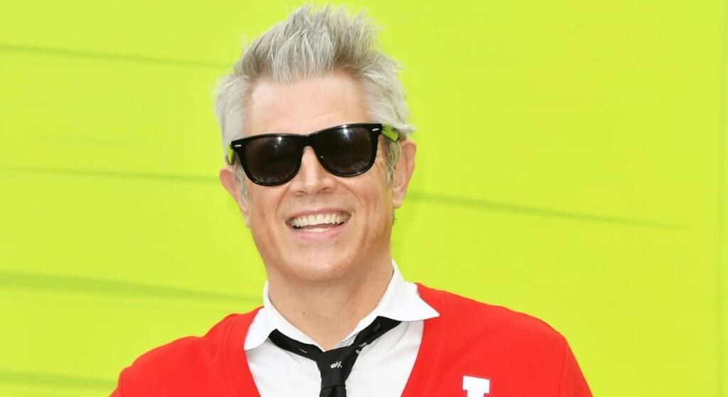 Johnny Knoxville wearing a read sweater and black tie.