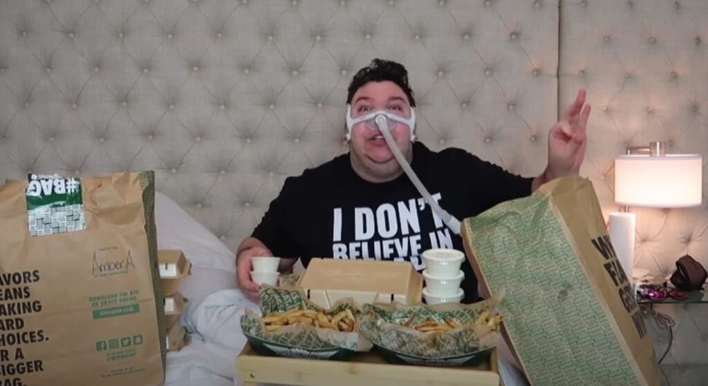 Nikocado Avacado on a bed with oxygen mask and bags of take out food.