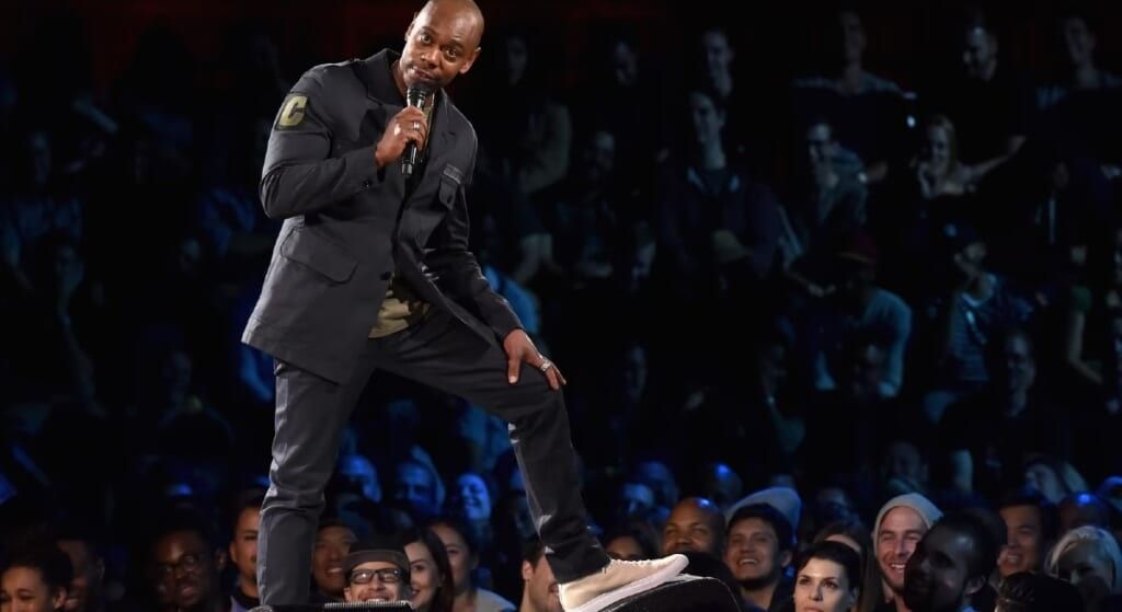 Dave Chappelle in all black suit holding a microphone during his standup.