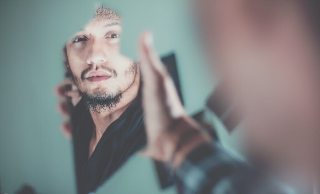 young man with facial hair looks in broken mirror
