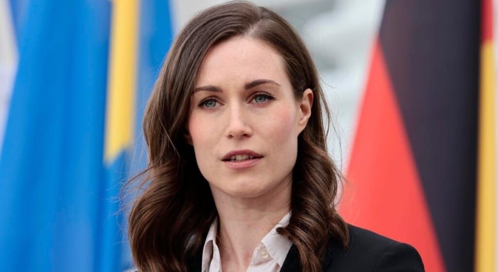 Prime Minister Sanna Marin in black suit standing in front of a flag.