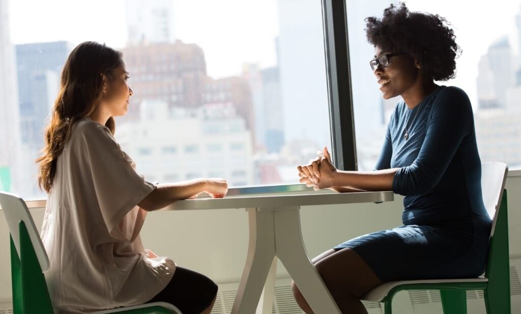 two women sit at table by the window conversing