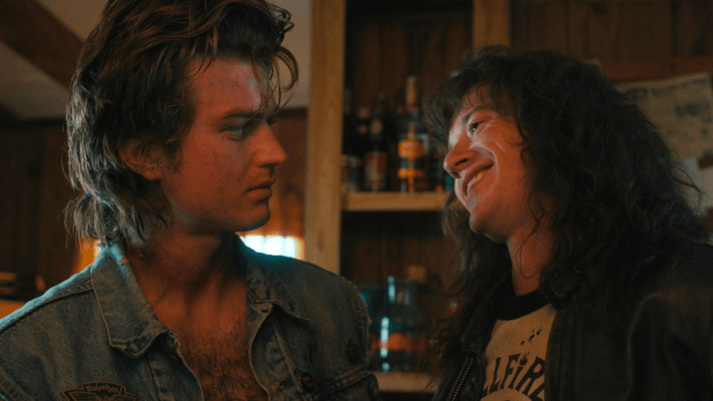 Steve Harrington turning to look at Eddie Munson, who is smiling cheekily at him.