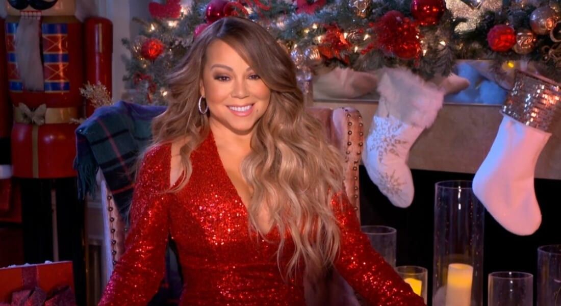 Mariah Carey sitting in front of a Christmas fireplace wearing a red dress.