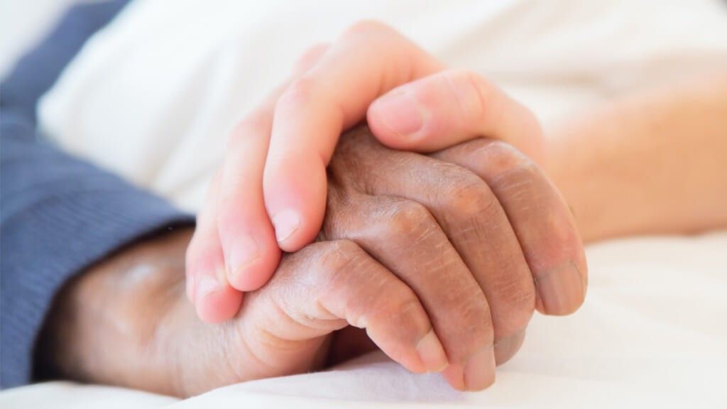 A white hand holding a black persons hand in a hospital bed 1024x576