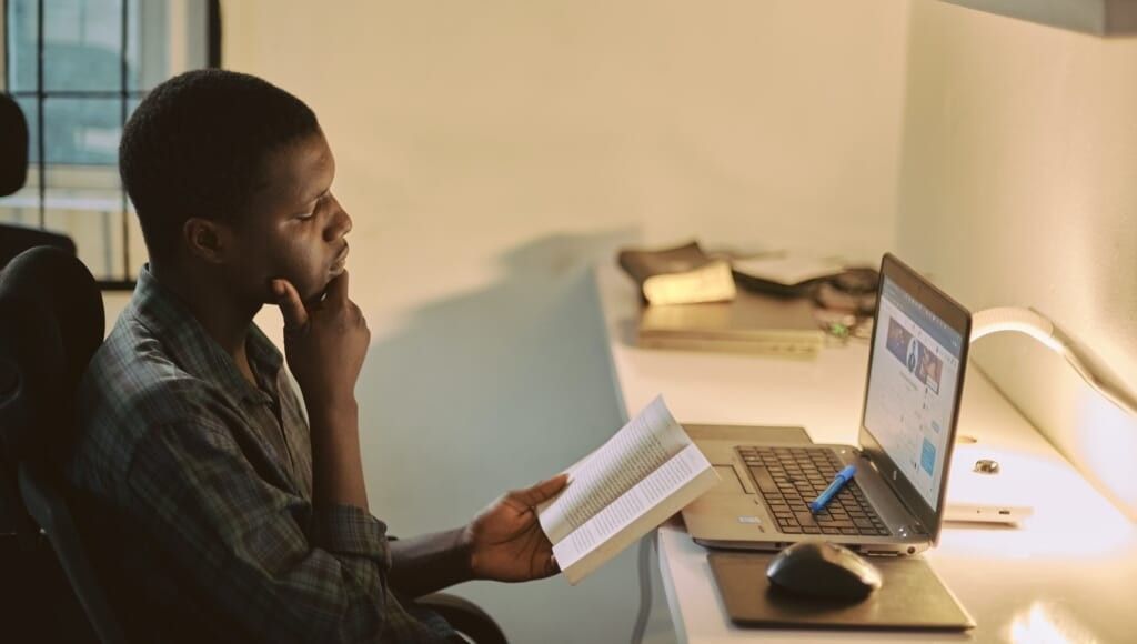 adult reads book sitting at desk in front of laptop