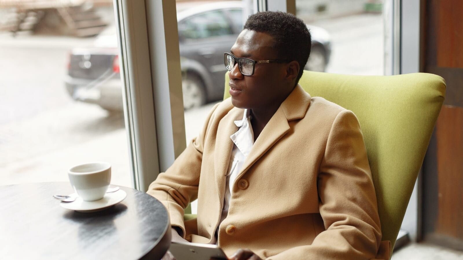 man in a light brown jacket staring out of a window with coffee before him